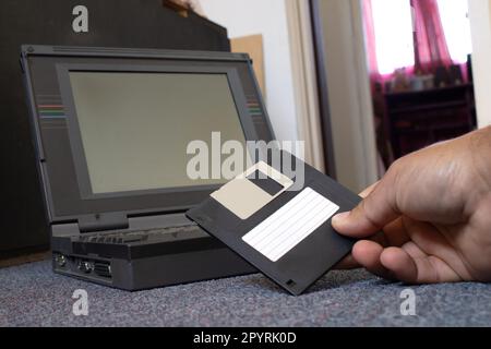 mains d'une personne prenant une disquette et la plaçant dans un vieil ordinateur portable. La personne est sur le sol d'une pièce Banque D'Images