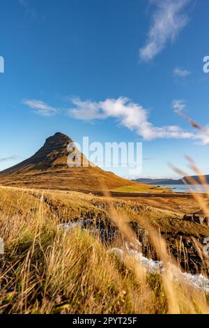 Vue sur la montagne Kirkjufell en Islande sur des champs de foin doré qui cachent un ruisseau d'eau et un ciel clair en arrière-plan Banque D'Images