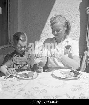 Une jeune fille et un garçon assis à l'extérieur à une table au soleil, la fille met quelque chose avec la fourchette et le couteau d'elle sur son assiette, le garçon, aveuglé par le soleil, fait une grimace. Banque D'Images