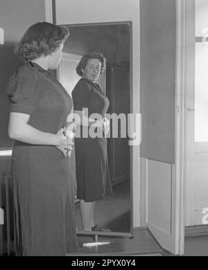 Une femme en robe se tient latéralement devant un miroir et se regarde pendant que ses mains couvrent son ventre. Banque D'Images