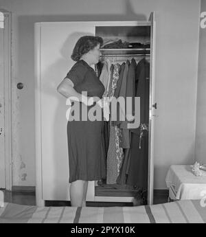 Une femme se tient devant une armoire ouverte dans une chambre à coucher et se examine dans le miroir de la porte du placard, tout en touchant sa taille avec ses mains. Banque D'Images