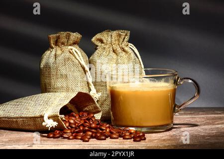 Sacs de grains de café torréfiés crus et une tasse de café en verre avec un reflet d'un store de fenêtre en arrière-plan Banque D'Images