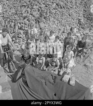 Photo non datée d'un groupe d'enfants regardant un spectacle de marionnettes parmi des piles de briques restantes des ruines. Les briques servent de rideau du théâtre de marionnettes (ci-dessous à gauche voilé par des couvertures). En arrière-plan, plus de piles de briques. À gauche, un adolescent est assis sur un vélo. Banque D'Images