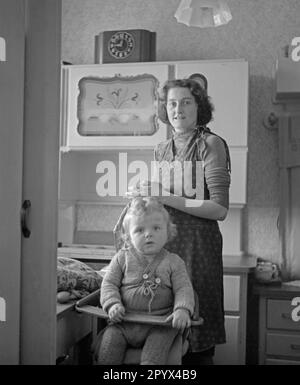 Photo sans date d'une veuve d'un soldat allemand tué dans la guerre prise dans une cuisine à manger à Berlin, probablement Berlin-Ouest, 1950. Au premier plan, son enfant s'assoit sur une chaise d'enfant. La femme porte une robe de tablier. Banque D'Images