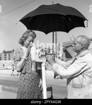 Photo non datée d'une jeune femme de Berlin-Ouest debout sur une balance mécanique, tout en étant pesée par un homme âgé dans une grenouille, qui fixe la balance et tient un parasol. La dame semble être surprise par le poids affiché. En arrière-plan, une rue, une jeune fille et des montagnes de décombres. Banque D'Images