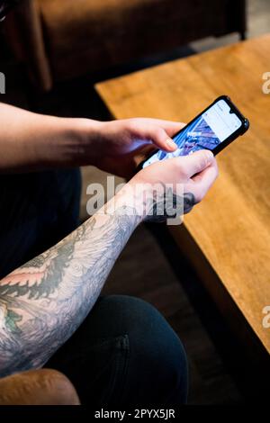 Mains tatouées tenant un smartphone défilant sur les médias sociaux assis sur un canapé avec une table basse devant la personne Banque D'Images