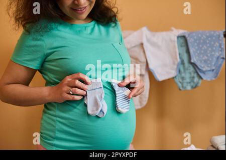 Gros plan d'une femme enceinte tenant des chaussettes bleues sur le ventre, debout contre des bodys repassés pour son nouveau-né Banque D'Images