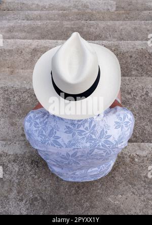 Un homme portant une chemise bleue et un grand chapeau de Panama beige sur sa tête, assis sur quelques marches de pierre et avec son dos à la caméra. Cadix, Espagne Banque D'Images