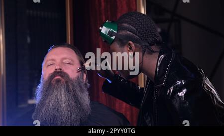 Une styliste afro-américaine en imperméable en cuir noir ravente la barbe d'un client dans un salon de coiffure Banque D'Images