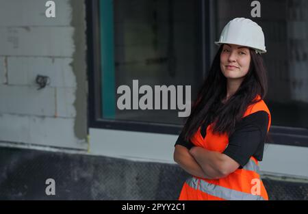 Jeune femme en casque blanc et gilet orange haute visibilité, cheveux longs foncés, regardant dans l'appareil photo, mains croisées confiance. Site de construction flou Banque D'Images