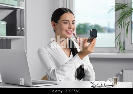 Jeune femme utilisant un miroir de poche cosmétique tout en appliquant le maquillage dans le bureau Banque D'Images