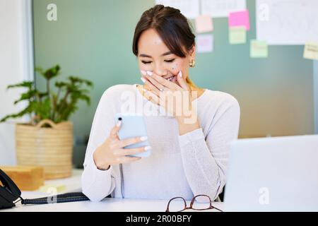 C'est un texte aussi peu flattant. une jeune femme d'affaires attirante assise seule au bureau et en train de rire tout en utilisant son téléphone portable. Banque D'Images