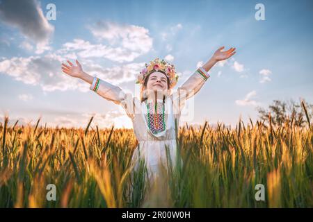 Belle jeune fille avec un chapelet de fleurs, robe folklorique ethnique avec broderie bulgare traditionnelle pendant le coucher du soleil sur un champ agricole de blé Banque D'Images