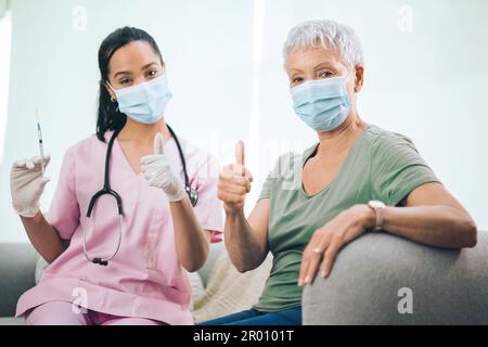 Plus qu'un médecin... Portrait d'un médecin masqué et de sa patiente masquée montrant les pouces vers le haut après avoir reçu une injection à la maison. Banque D'Images