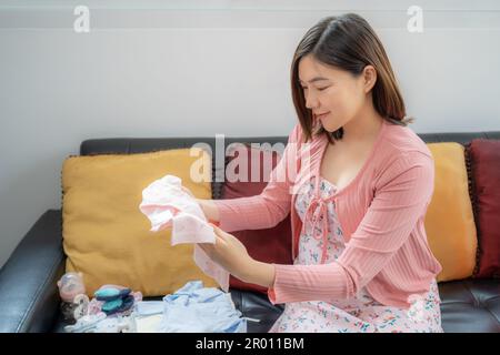 Une jeune femme asiatique heureuse, vêtue de vêtements de maternité, souriant et regardant un nouveau vêtement de bébé qu'elle tient tout en étant assise dans sa maison lumineuse Banque D'Images