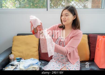 Une jeune femme asiatique heureuse, vêtue de vêtements de maternité, souriant et regardant un nouveau vêtement de bébé qu'elle tient tout en étant assise dans sa maison lumineuse Banque D'Images