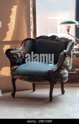 Fauteuil classique vintage avec de beaux éléments en bois sculpté et une garniture bleue déchirée dans la salle de grunge avec lampe verte sur le seuil de la fenêtre. Concept intérieur rétro avec espace de copie. Banque D'Images