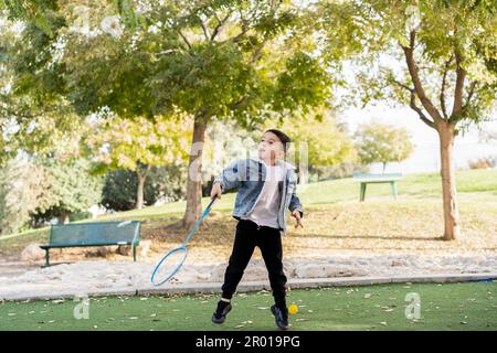 Kid Jumping et frapper shuttlecock avec raquette. Enfant actif jouant au badminton dans le parc de Sunny City, s'amuser avec la raquette et la navette sur le terrain de Green Grass Field, nature Background. Banque D'Images