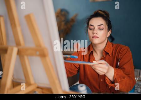 Une artiste brésilienne concentrée avec de la peinture sur ses mains travaille sur le dessin d'une image sur toile assise devant elle, appliquant un coup de pinceau sur Banque D'Images