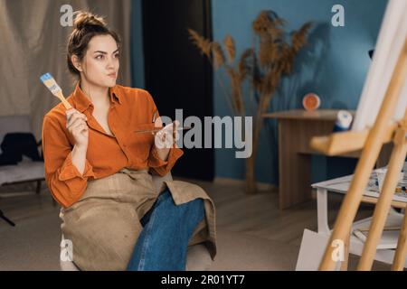 Femme peintre d'art ayant une expression réfléchie assis avant son chevalet. Crise créative. Peintre créant des œuvres d'art pour la galerie d'images Banque D'Images