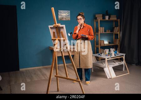 femme latino concentrée inspirante en tablier dessine des images avec des peintures, debout dans un studio moderne ou salon. l'artiste féminine pensive regarde Banque D'Images