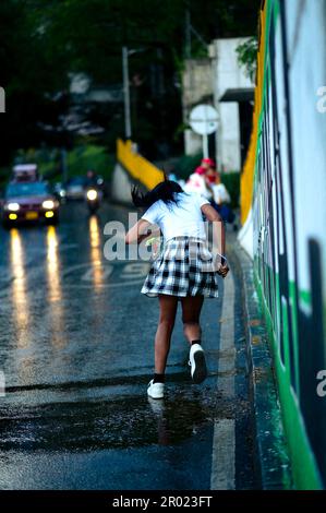 Femme en train de courir dans la rue sous la pluie, jeune femme en jupe à carreaux et chemise blanche en courant pour éviter la pluie soudaine tenant le smartphone Banque D'Images