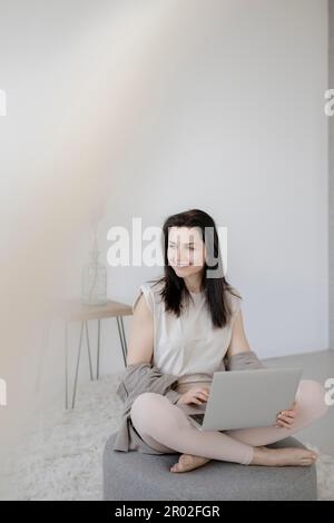 Jeune femme assise confortablement avec un ordinateur portable sur un tabouret dans le salon Banque D'Images