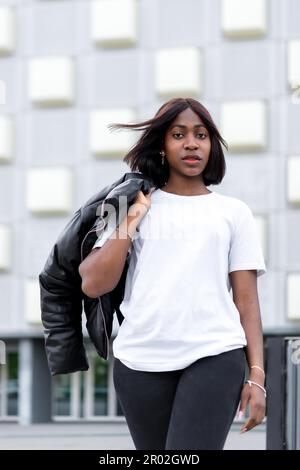 Une ville à la mode : une jeune femme noire pose dans un t-shirt blanc élégant et un pantalon noir, avec un bâtiment gris comme toile de fond urbaine chic Banque D'Images