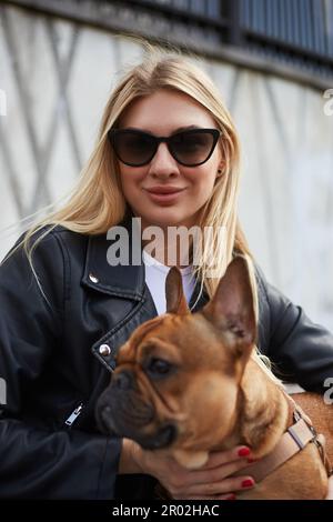 Belle femme blonde dans des lunettes de soleil posant sur un banc avec un bulldog français sur ses tours. Joyeux amoureux de chien qui apprécie la journée Banque D'Images