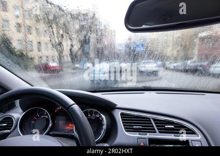 Le tableau de bord d'un intérieur de voiture avec vue sur les silhouettes des bâtiments d'une rue de ville floue par la pluie sur le pare-brise. Banque D'Images