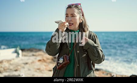 Femme hispanique mature avec cheveux gris touriste portant sac à dos parlant au téléphone au bord de la mer Banque D'Images