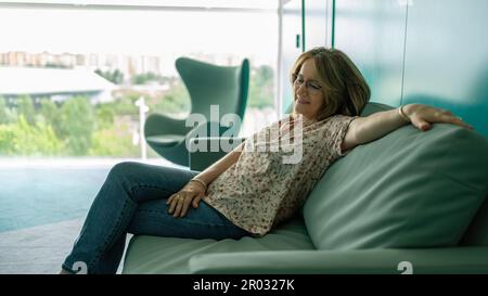 Femme assise dans une posture détendue sur un canapé et souriante dans une attitude heureuse et calme. Banque D'Images