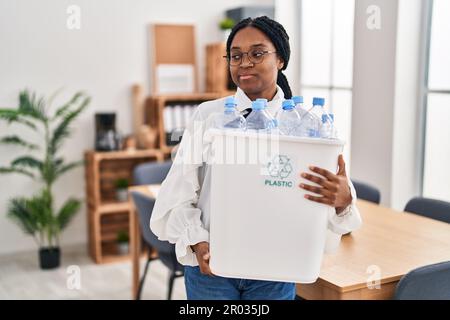 Femme afro-américaine travaillant au bureau tenant une bouteille en plastique pour le recyclage souriant regardant à côté et regardant loin de penser. Banque D'Images