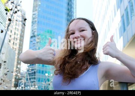 Belle jeune fille joyeuse d'adolescence dans un haut de lavande sur le fond de la ville montre les pouces vers le haut la publicité heureuse souriant toutes les dents Voyage Banque D'Images