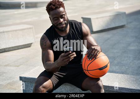 S'adapte au jeune sportif noir tenant le ballon de basket-ball en utilisant le téléphone à l'extérieur. Banque D'Images