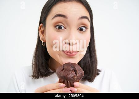 Gros plan d'une femme asiatique mignonne, tenant un cupcake au chocolat près de la bouche, en mordant, aime manger des pâtisseries, fond blanc Banque D'Images