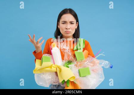 Une jeune femme en colère clenches poing, retient les déchets plastiques non triés, recycle, semble agacé, fond bleu Banque D'Images
