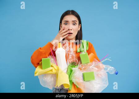 Choqué femme asiatique, semble embarrassée à quelque chose d'horrible, tenant des déchets en plastique, triant les déchets pour le recyclage, fond bleu Banque D'Images