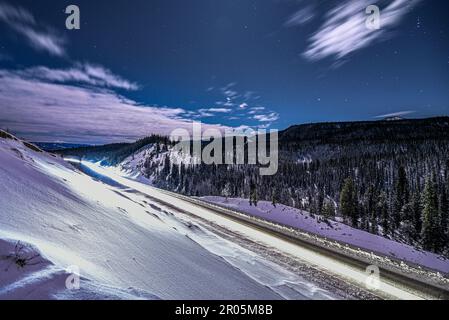 Scène nocturne d'une route d'hiver dans le nord du Canada, territoire du Yukon. Prise sur la nuit de lune éclairée avec ciel étoilé bleu vif, épinette, arbres entourant. Banque D'Images