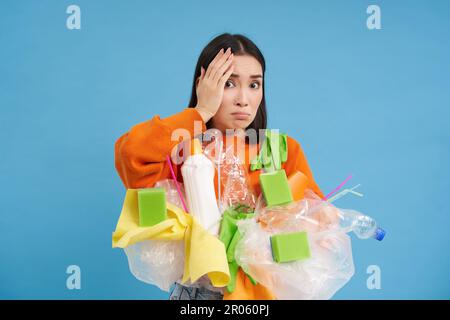 Choqué femme asiatique, semble embarrassée à quelque chose d'horrible, tenant des déchets en plastique, triant les déchets pour le recyclage, fond bleu Banque D'Images