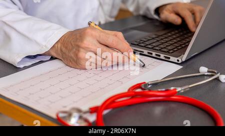 cardiologue médecin analyse l'électrocardiogramme, bureau avec stéthoscope rouge sur la table et ordinateur portable en arrière-plan Banque D'Images