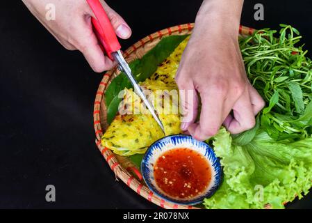 Mains avec des ciseaux coupant crêpes vietnamiennes Banh xeo avec de la nourriture de mer sur fond noir vue de dessus Banque D'Images