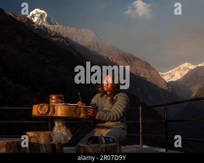 Brunette Backpacker sur son chemin au camp de base d'Annapurna écrire les expériences de son jour sur son laiterie avec l'Himalaya en arrière-plan. Banque D'Images