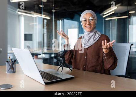 Une jeune femme musulmane dans un hijab est assise dans un bureau à un bureau devant un ordinateur portable et un microphone. parle lors d'un appel vidéo, présente un projet. Gestuelle avec ses mains, souriant à la caméra. Banque D'Images
