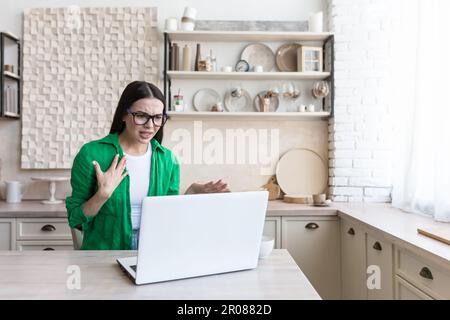 Portrait d'une femme contrariée assise à la maison dans la cuisine et parlant malheureuse par un appel vidéo. Argumenter, discuter, indigner. Banque D'Images