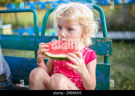 Une petite fille mange de la pastèque en été assise dans le pays sur un vieux banc. Bonne enfance. Alimentation saine pour les enfants. Banque D'Images