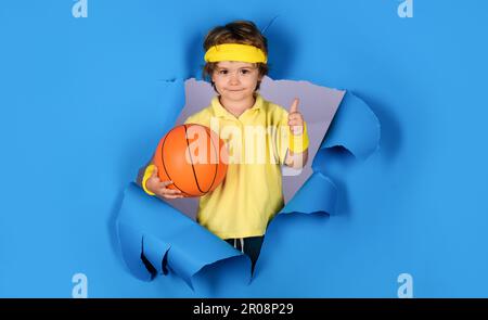 Jeu d'entraînement de basket-ball. Un enfant souriant avec une balle de basket-ball montrant le pouce vers le haut. Profitez d'un jeu sportif. Mignon garçon jouant au basket-ball. Petit ballon de base Banque D'Images