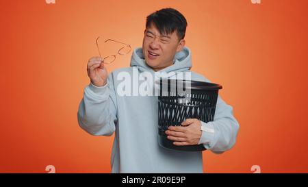 Homme asiatique qui se déporte, qui jette des lunettes dans la poubelle après une chirurgie de traitement par laser de vision médicale, en regardant sourire la caméra. Un charmant Chinois isolé seul sur fond orange studio Banque D'Images