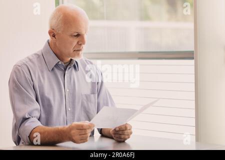 un homme d'âge moyen avec un morceau de papier dans sa main , assis à une table dans une installation moderne remplie de lumière. Il peut être en attente d'un service médical ou Banque D'Images