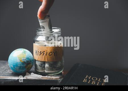 Sauver des bocaux pleins d'argent et de globe avec la Sainte Bible pour la mission, Mission christian idée. Main tenant dollar avec la bible sur table en bois, bac chrétien Banque D'Images